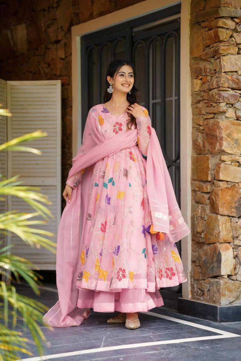 Woman in a pink traditional outfit with floral patterns standing in front of a stone wall. | 5012 Beautiful Star Georgette Embroidery Floral Worked Anarkali Kurti Collection