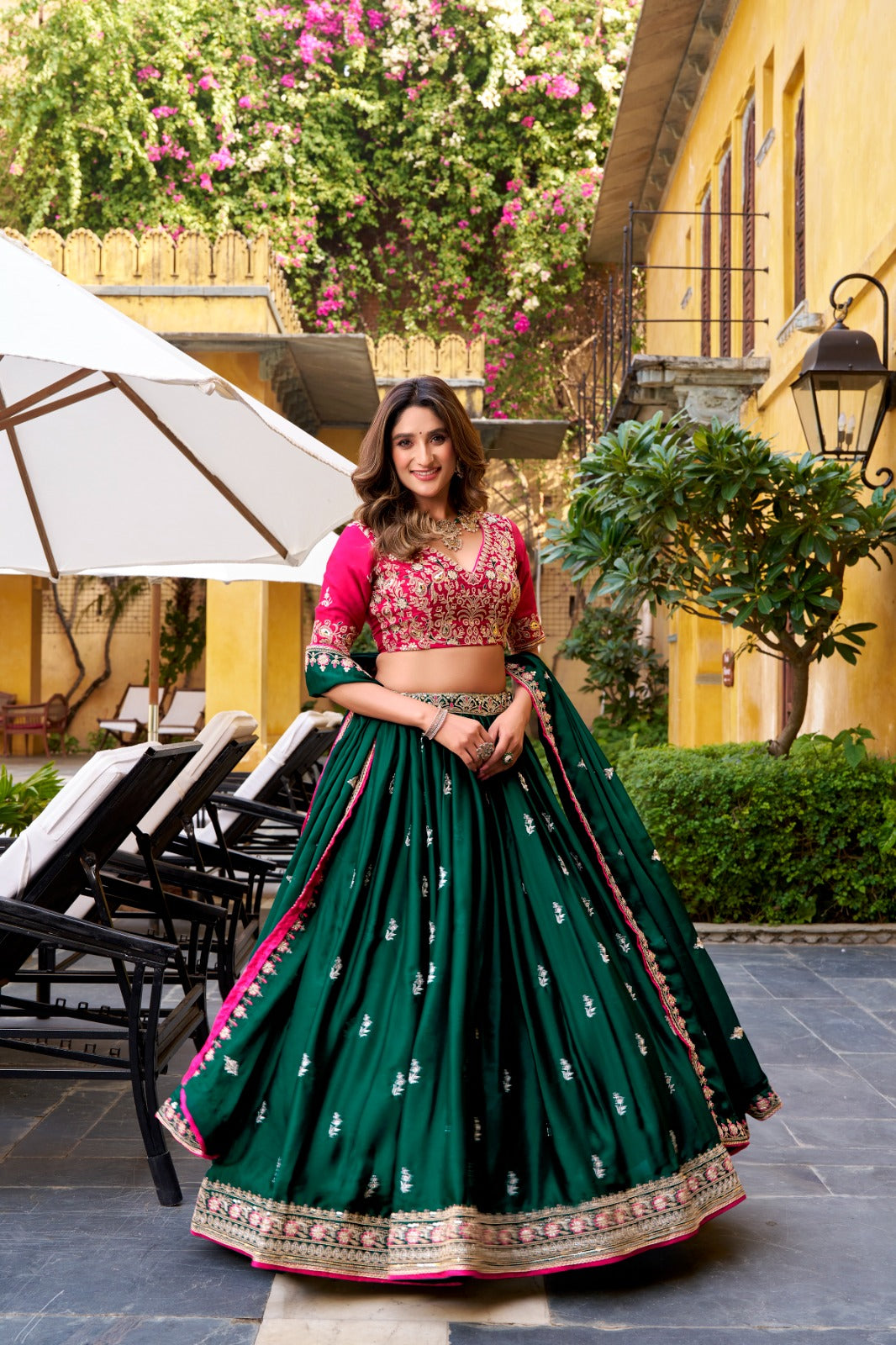 Woman in a green and pink traditional outfit standing outdoors with a building and plants in the background. | Amrah Wholesale Taraa L 1449 Silk Satin Sequins Worked Designer Lehenga Choli Wholesale Price