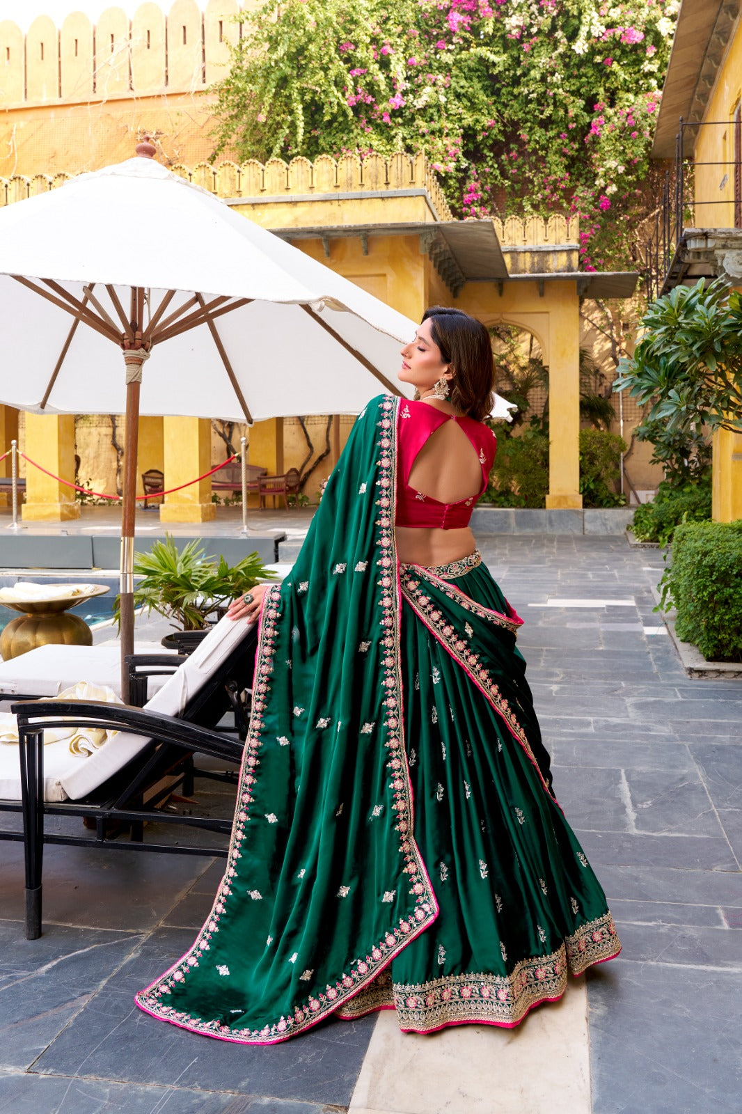 Woman in a green saree with a red blouse standing outdoors with an umbrella and garden in the background. | Amrah Wholesale Taraa L 1449 Silk Satin Sequins Worked Designer Lehenga Choli Wholesale Price