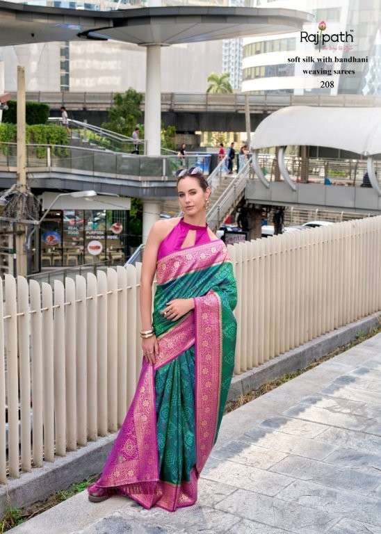 Woman in a green and pink saree standing on a sidewalk with a modern building in the background | RAJPATH Sugandh Silk Pure Silk Bandhani Sarees Catalogue